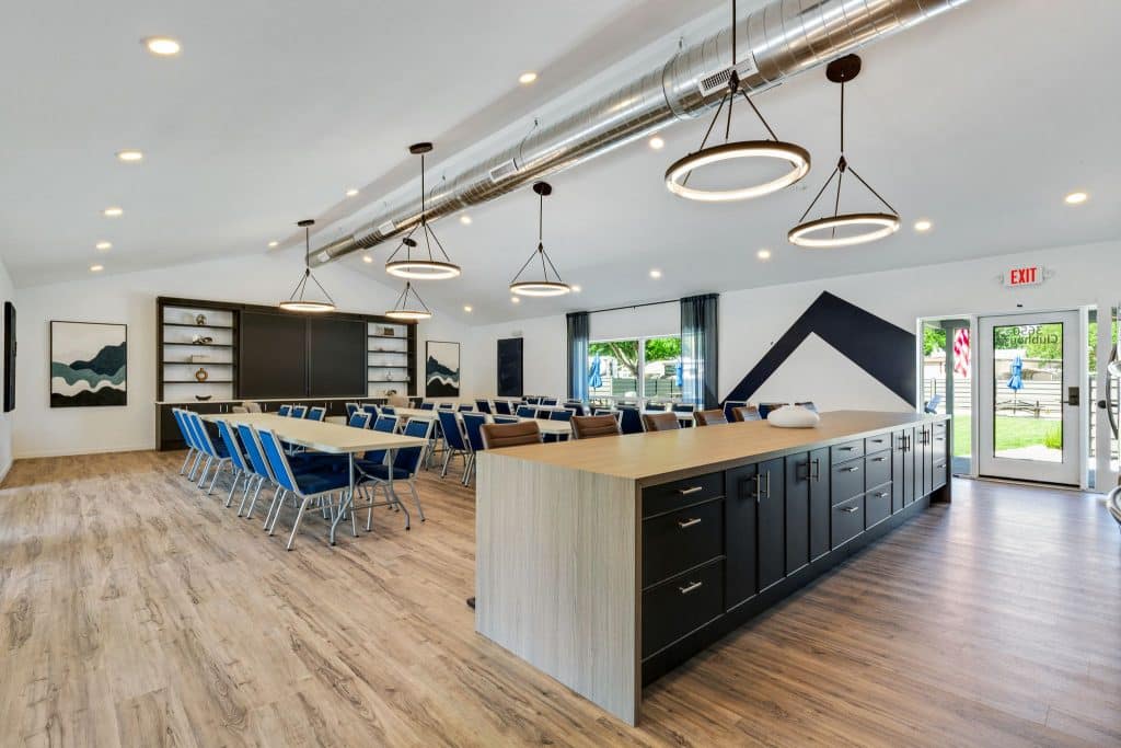 A spacious community room with long tables, blue chairs, a black island countertop, pendant lighting, and access to an outdoor patio in englewood co