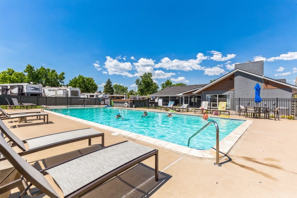A fenced outdoor pool with people swimming, surrounded by lounge chairs and umbrellas, adjacent to a gray building and RVs in the background in englewood co